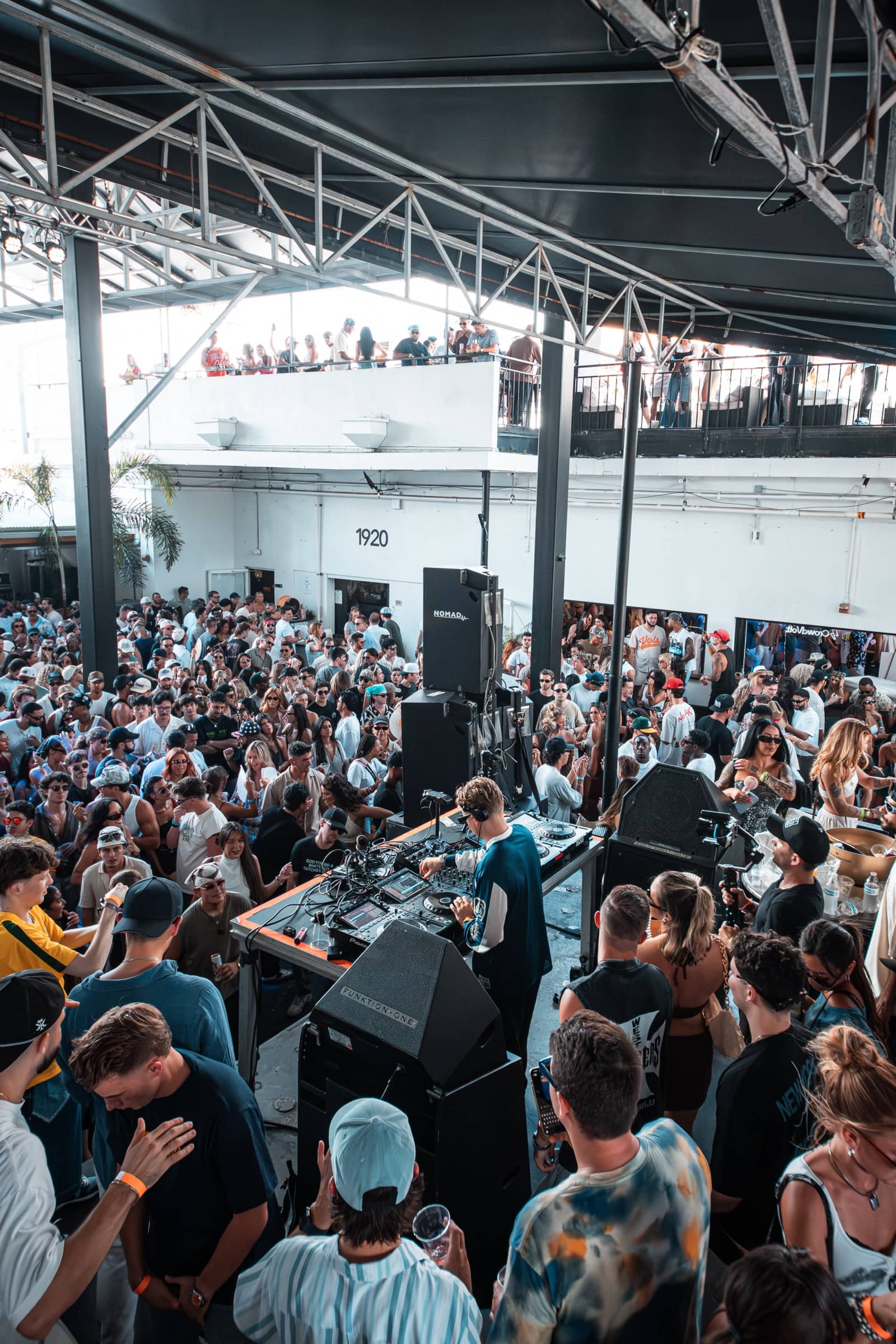Open-air warehouse packed with a daytime crowd, DJ playing under steel roof trusses