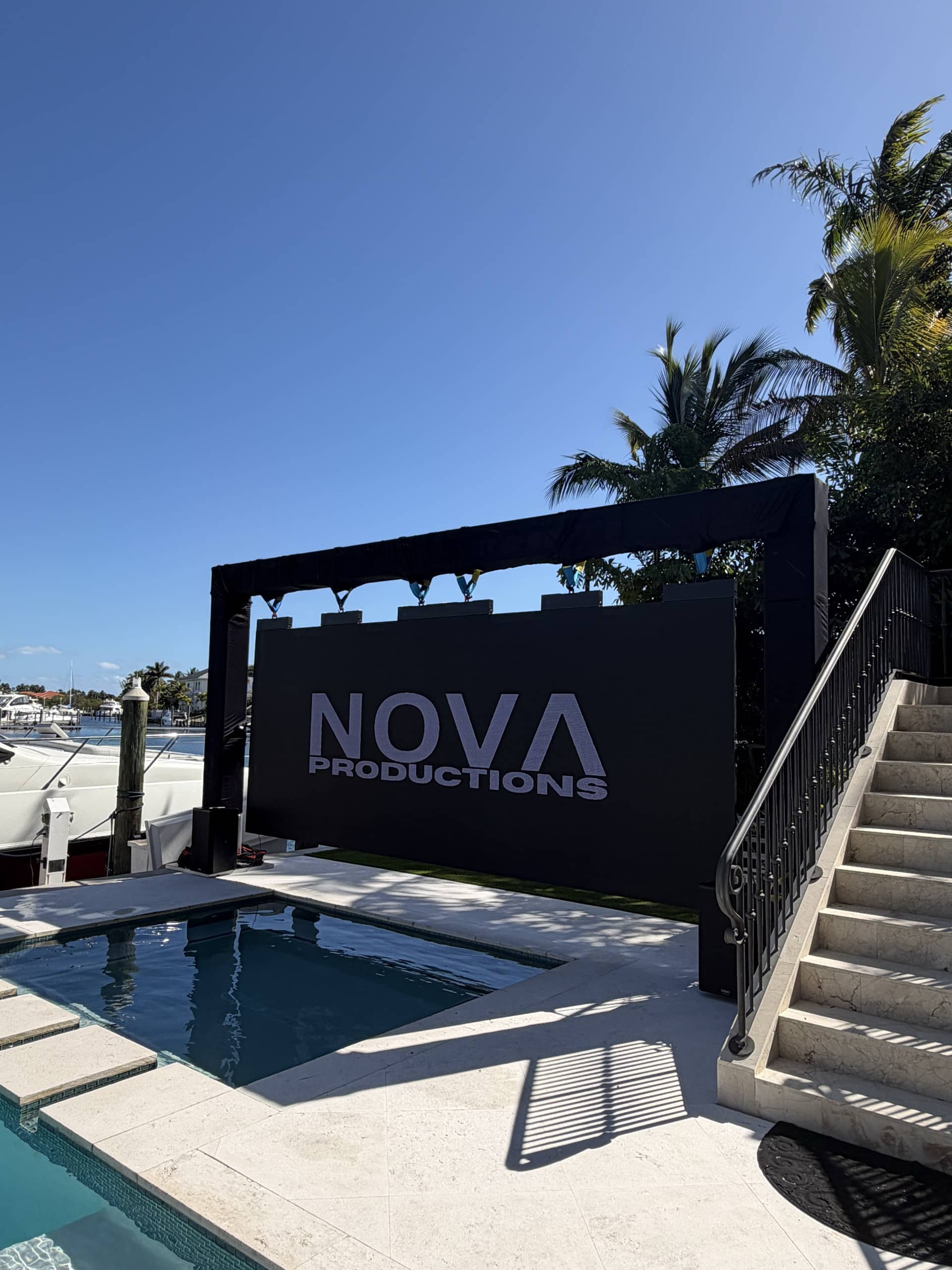 Outdoor LED wall over a pool showing the Nova Productions logo, with palms and the waterway behind